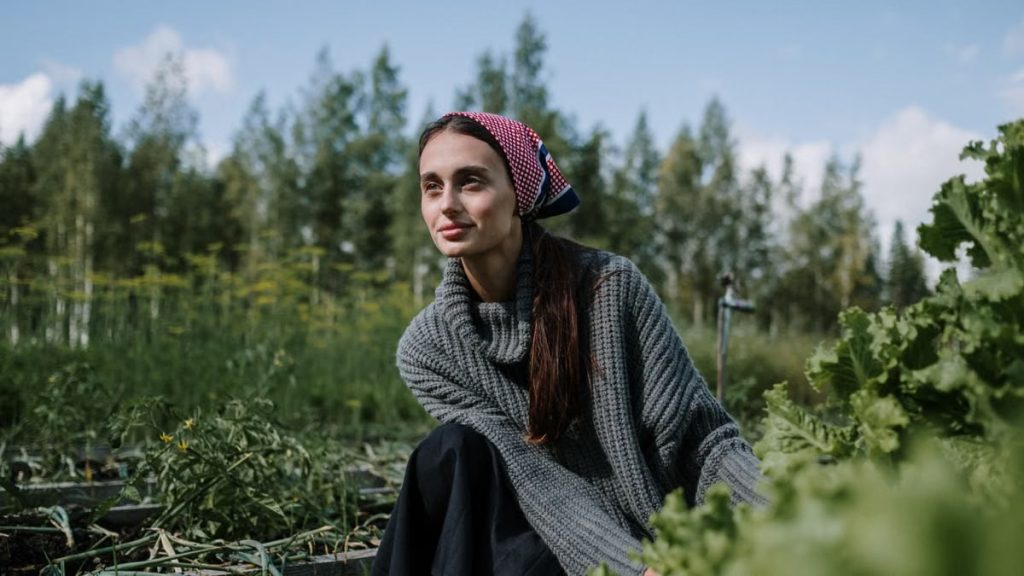 Woman in a headscarf tending to a garden in a lush green setting. Outdoors with clear sky.