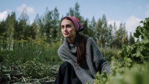 Woman in a headscarf tending to a garden in a lush green setting. Outdoors with clear sky.